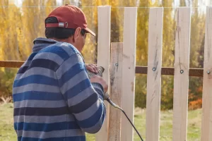 A man using a drill to tighten a screw while installing a picket fence, illustrating fence installation.