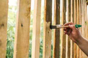 A hand applying brown wood stain to a wooden fence with greenery in the background, highlighting DIY repairs.